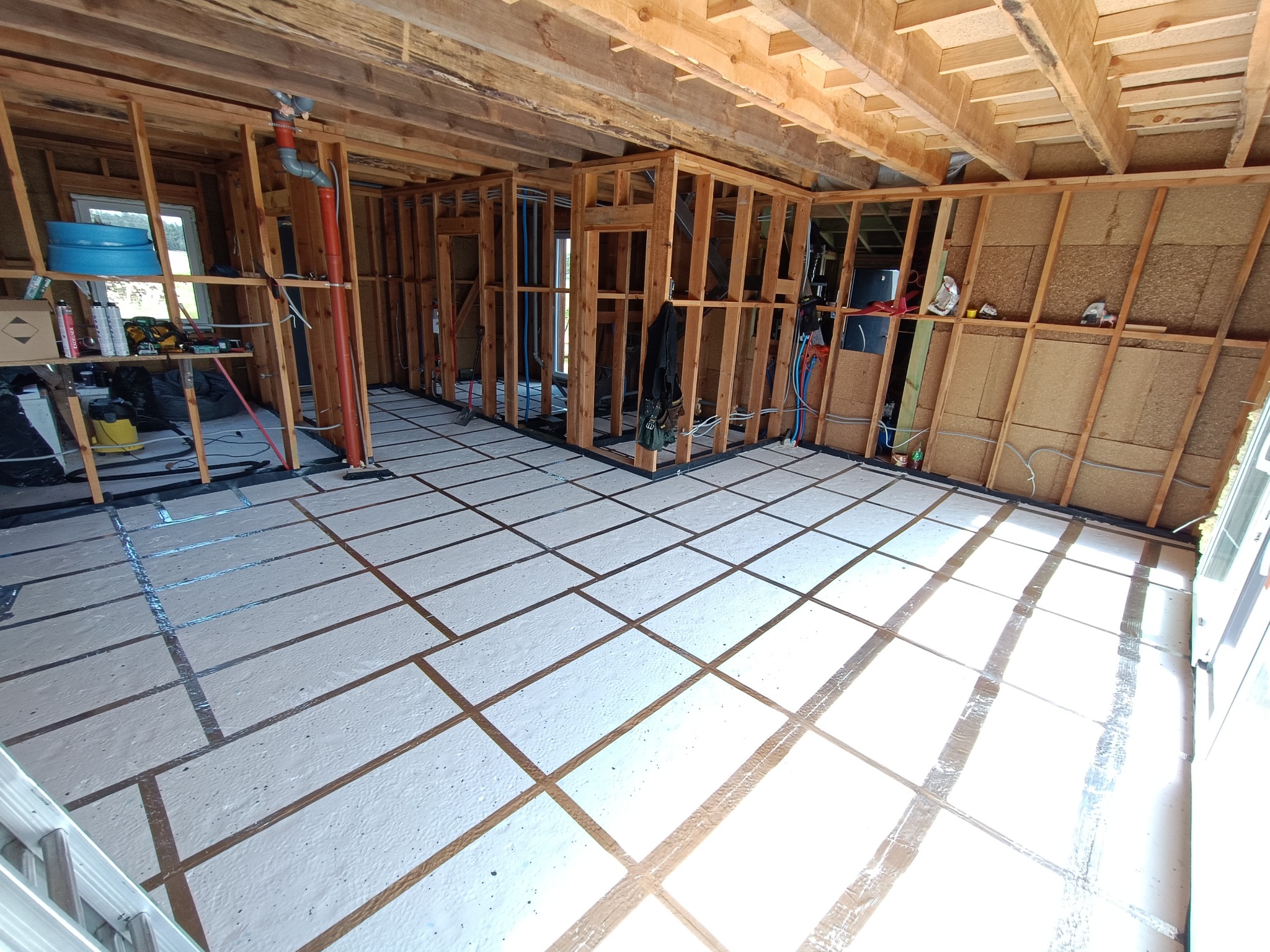 Wnętrze domu w trakcie budowy: drewniany szkielet, izolacja podłogi w kratkę, instalacje. Widoczne narzędzia i materiały budowlane. Jasne oświetlenie.