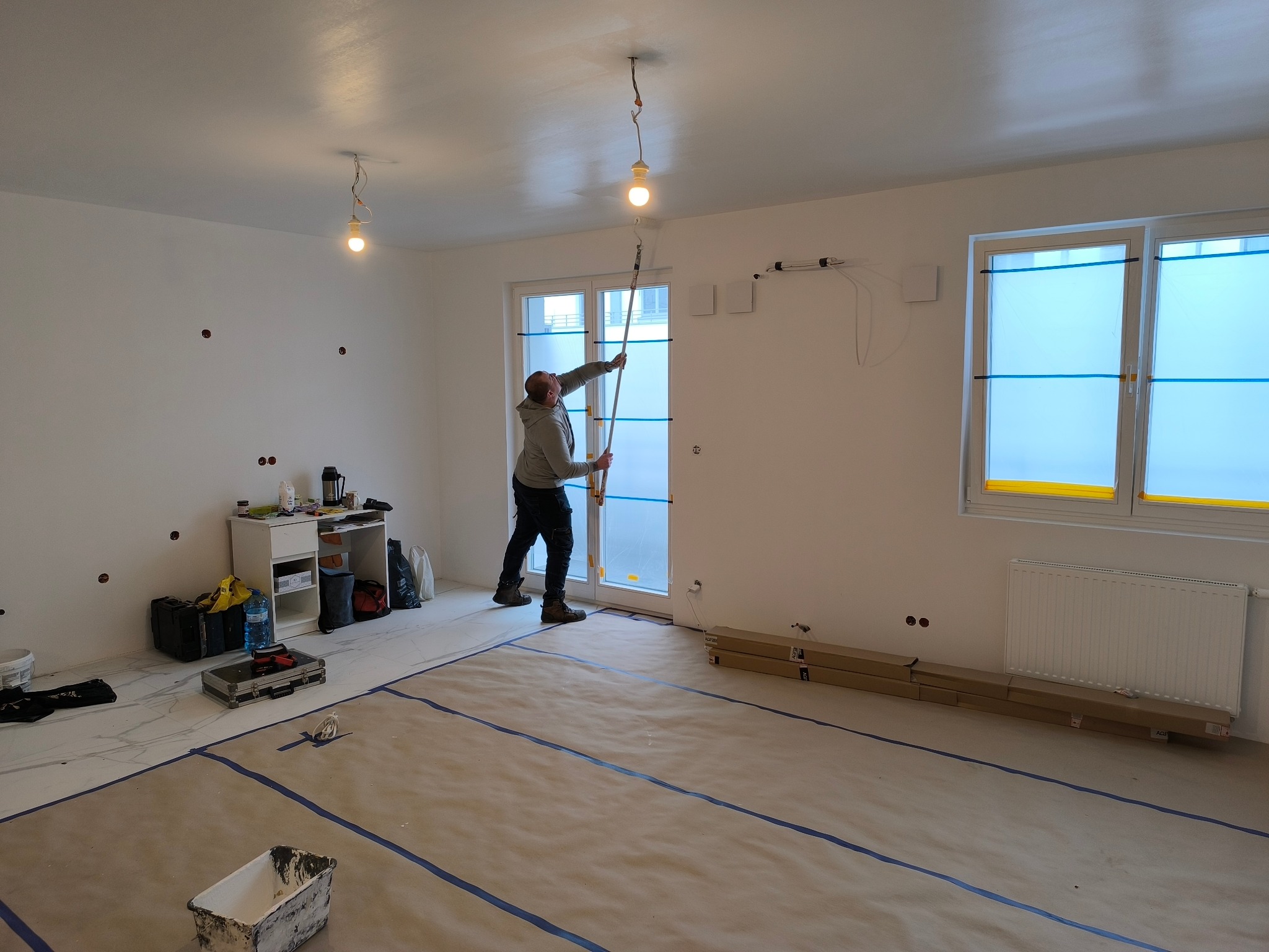 Malowanie sufitu w jasnym pokoju z zabezpieczoną podłogą. Widoczny malarz z wałkiem na teleskopie, narzędzia i osłonięte okna. Etap prac wykończeniowych.