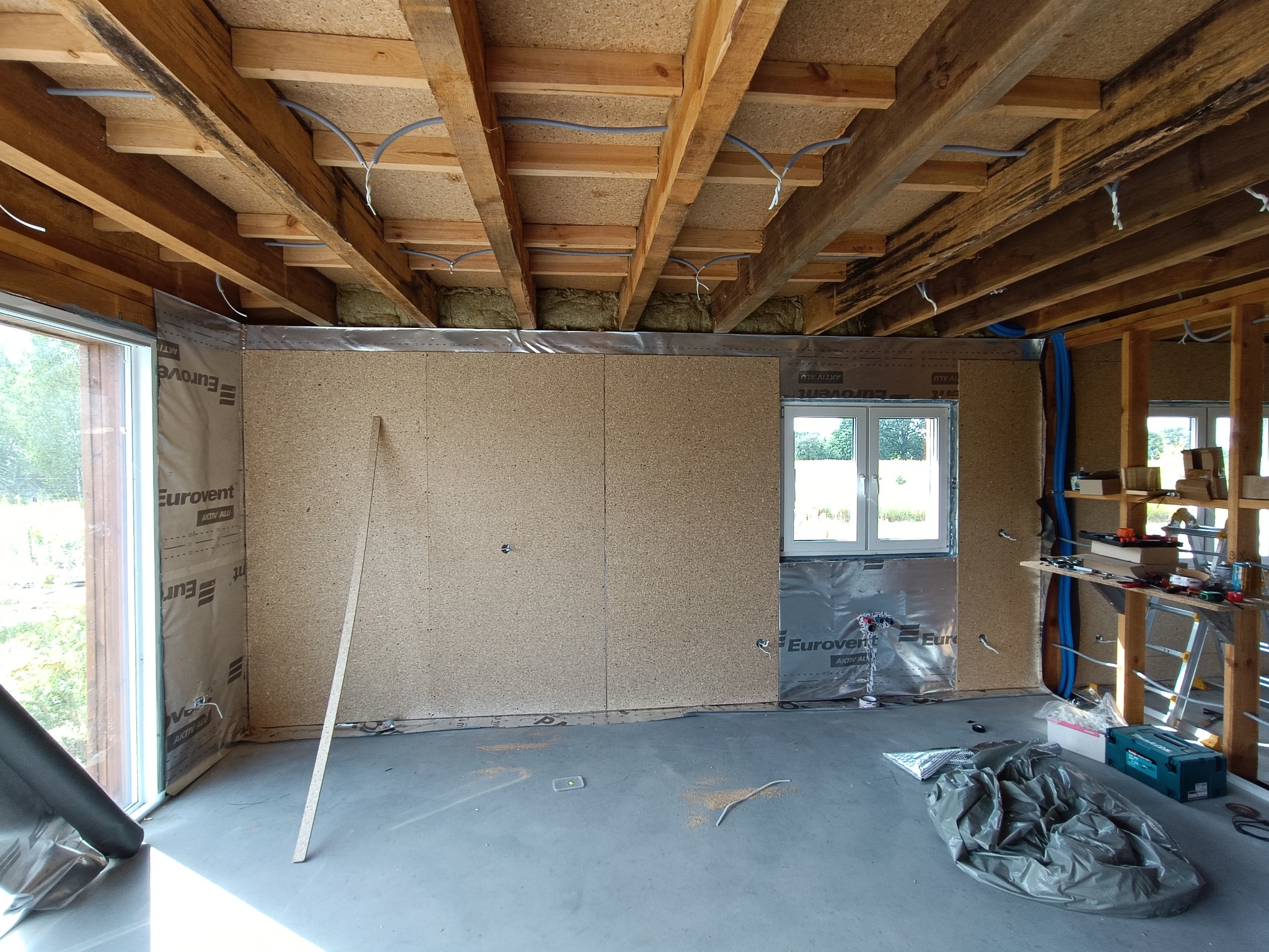 Wnętrze domu w trakcie budowy: ściany z płyty OSB, izolacja, drewniany strop z instalacjami. Widoczne okno i narzędzia. Surowy stan wykończenia.