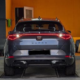 Wypolerowani Ernest Milewski - Tył szarego samochodu marki Cupra z włączonymi światłami, widoczny w warsztacie na tle pomarańczowej ściany.