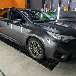 Wypolerowani Ernest Milewski - Szary samochód typu kombi na kratce w warsztacie, widoczne okna zakryte folią, oświetlenie warsztatowe i narzędzia na półce.