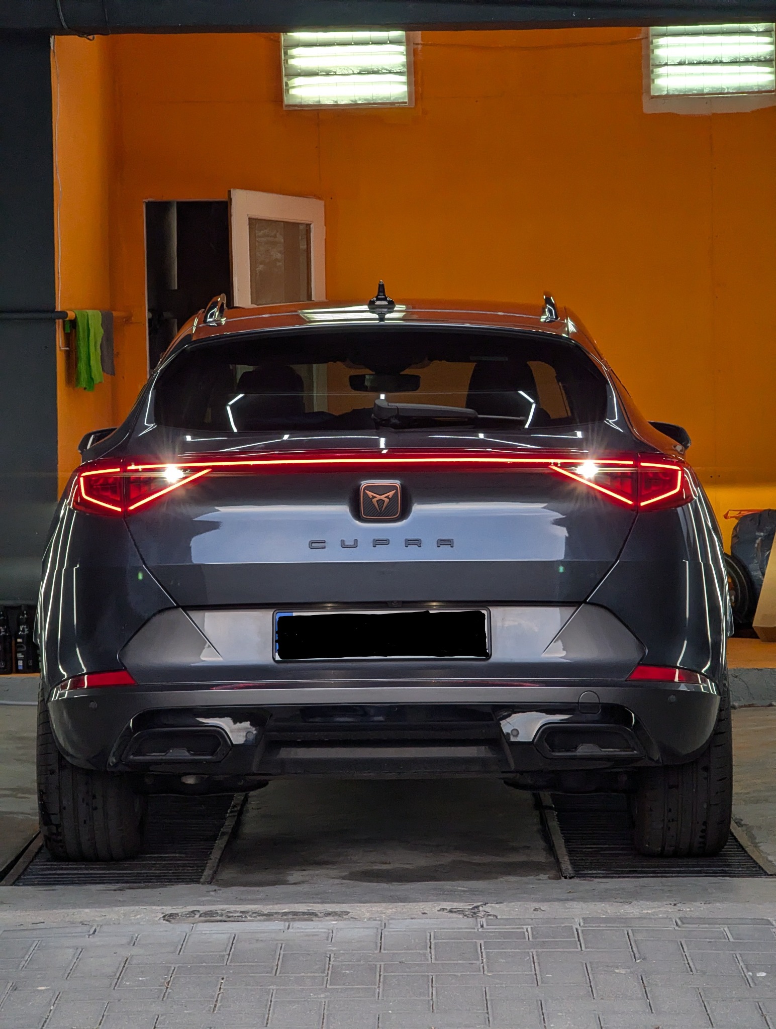 Tył szarego samochodu marki Cupra z włączonymi światłami, widoczny w warsztacie na tle pomarańczowej ściany.