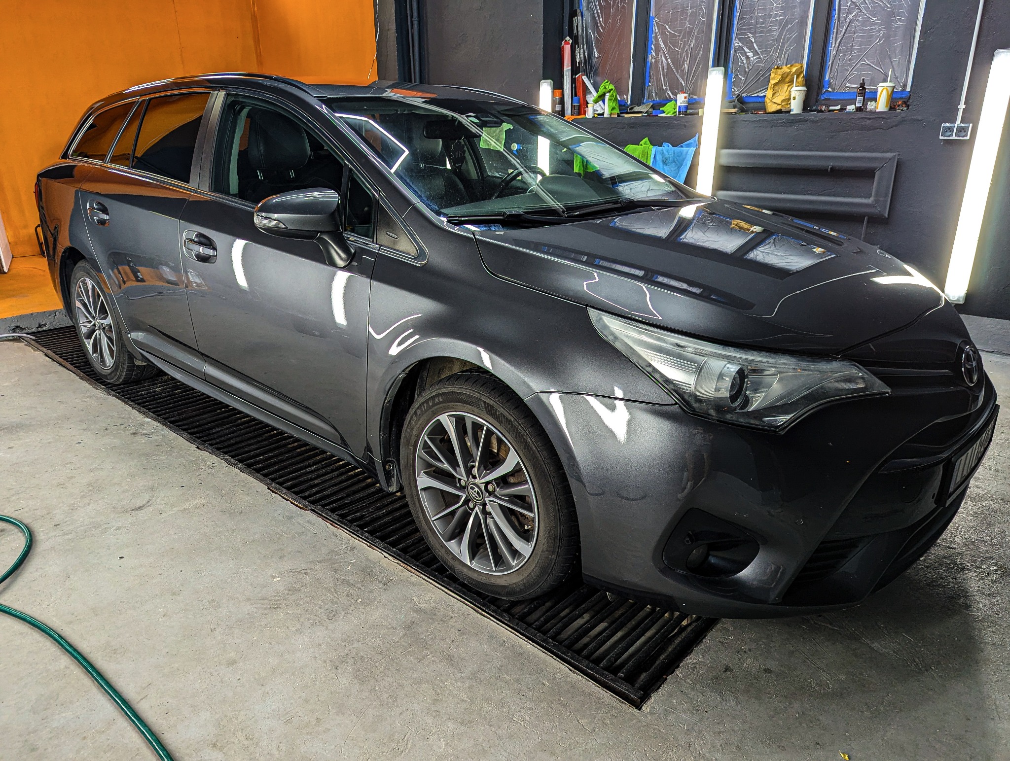 Szary samochód typu kombi na kratce w warsztacie, widoczne okna zakryte folią, oświetlenie warsztatowe i narzędzia na półce.