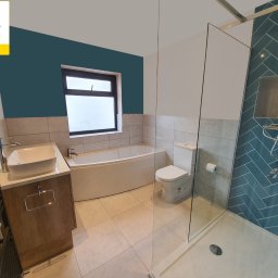 PROJEKT Pro. - Wykończona łazienka z wanną, toaletą, umywalką na szafce z drewnianym frontem oraz kabiną prysznicową z turkusowymi płytkami ułożonymi w jodełkę.