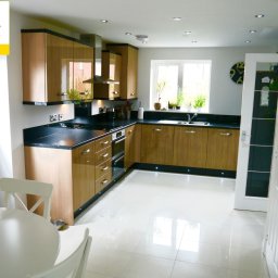 PROJEKT Pro. - Nowoczesna kuchnia z drewnianymi szafkami, czarnym blatem i błyszczącymi, białymi płytkami podłogowymi, widok z perspektywy stołu z białymi krzesłami. W oknie widoczne rośliny doniczkowe.