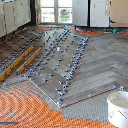 PROJEKT Pro. - Układanie płytek podłogowych w jodełkę w kuchni, widoczne poziomice, system poziomowania płytek z niebieskimi klipsami, pomarańczowa mata podkładowa, białe szafki kuchenne w tle.