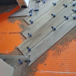 PROJEKT Pro. - Układanie podłogi z płytek imitujących drewno w jodełkę, z użyciem poziomicy i systemu poziomowania płytek na pomarańczowej macie izolacyjnej.