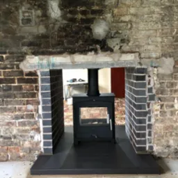 Wnęka kominkowa w trakcie przebudowy: surowe cegły po bokach kontrastują z nową, ciemną okładziną z płytek, w tle widoczny piec wolnostojący i wyposażenie pokoju.