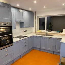 PROJEKT Pro. - Kuchenna zabudowa w odcieniach szarości z białym blatem, dwukomorowy zlew pod oknem, płyta indukcyjna i piekarnik w zabudowie, pomarańczowa podłoga, widoczne gniazdka elektryczne.