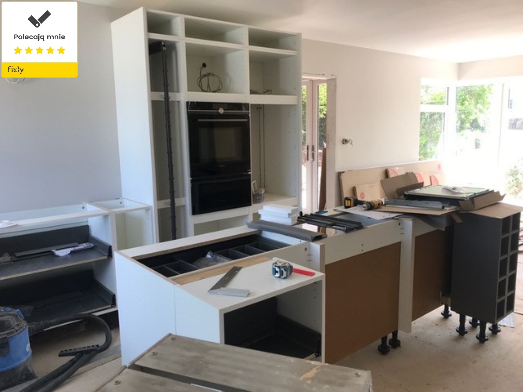 Montaż białej zabudowy kuchennej z wbudowanym piekarnikiem, widoczne narzędzia i akcesoria montażowe, częściowo zdemontowane elementy, okno w tle.