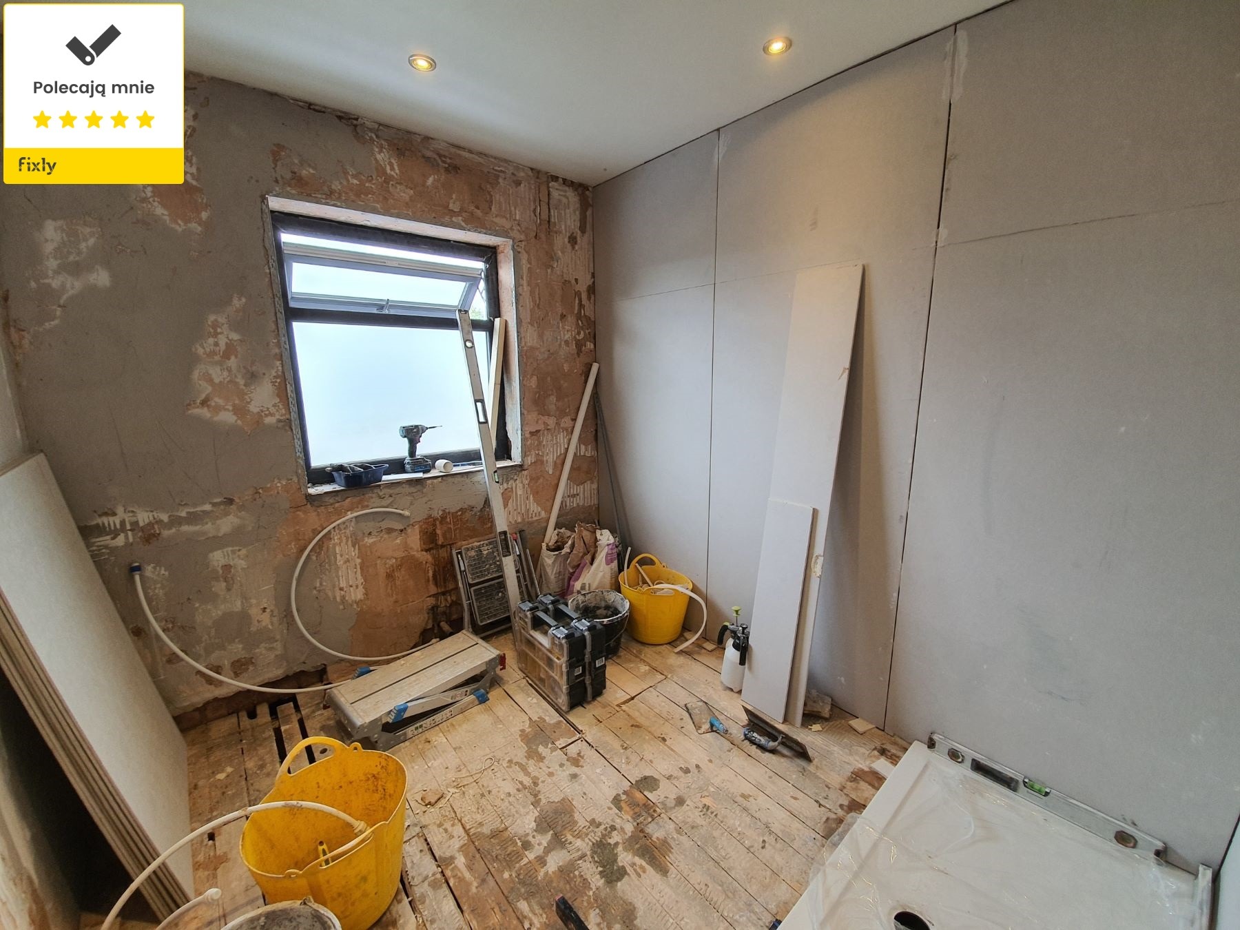 Remont łazienki w trakcie: ściany częściowo pokryte płytami gipsowo-kartonowymi, widoczne instalacje, narzędzia i materiały budowlane, podłoga drewniana z zabrudzeniami, okno z widocznym wiertłem.