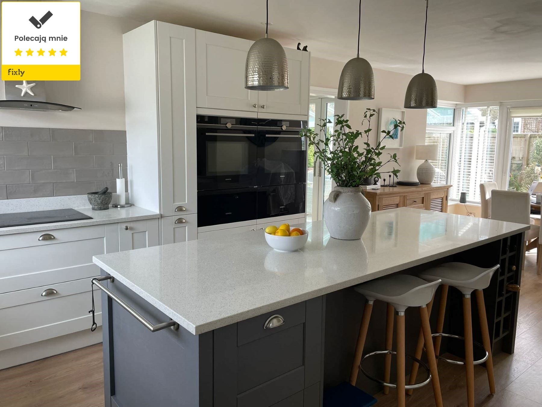 Nowoczesna kuchnia z wyspą, białymi szafkami z frezowanymi frontami, szarymi elementami i blatem z jasnego kamienia, oświetlona trzema metalowymi lampami wiszącymi.