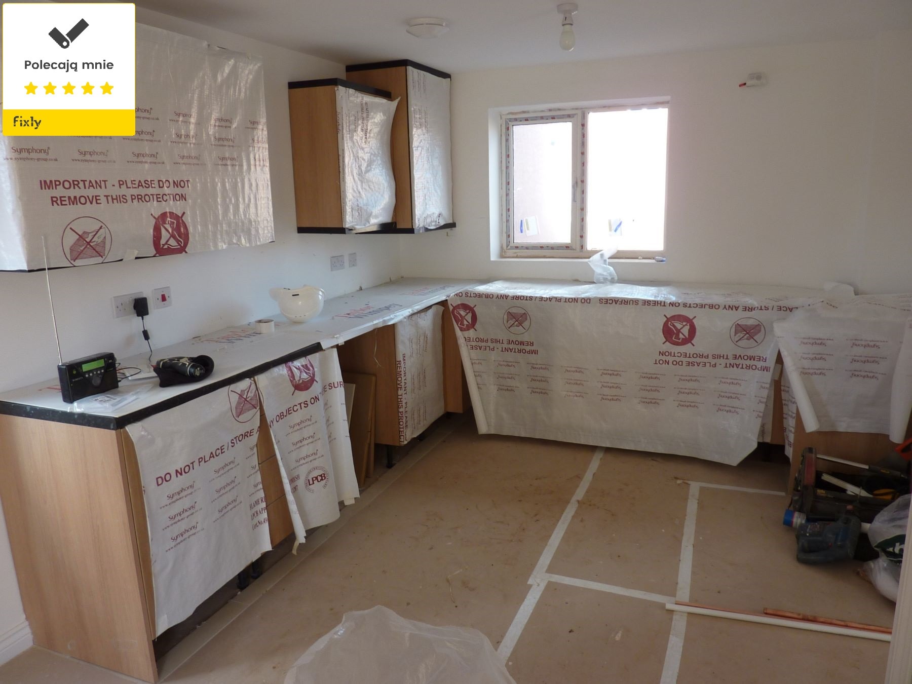 Kuchnia w trakcie zabezpieczania na czas prac remontowych; meble i blaty osłonięte folią ochronną z nadrukiem, widoczne narzędzia na podłodze, surowa podłoga przygotowana do dalszych prac...