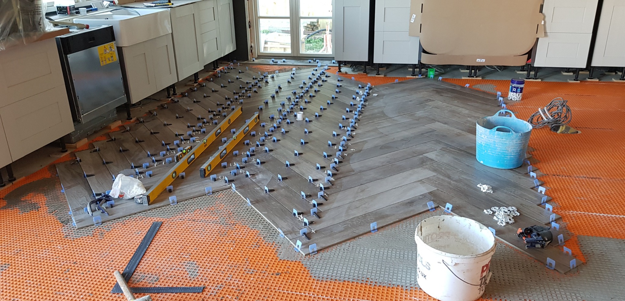 Układanie płytek podłogowych w jodełkę w kuchni, widoczne poziomice, system poziomowania płytek z niebieskimi klipsami, pomarańczowa mata podkładowa, białe szafki kuchenne w tle.