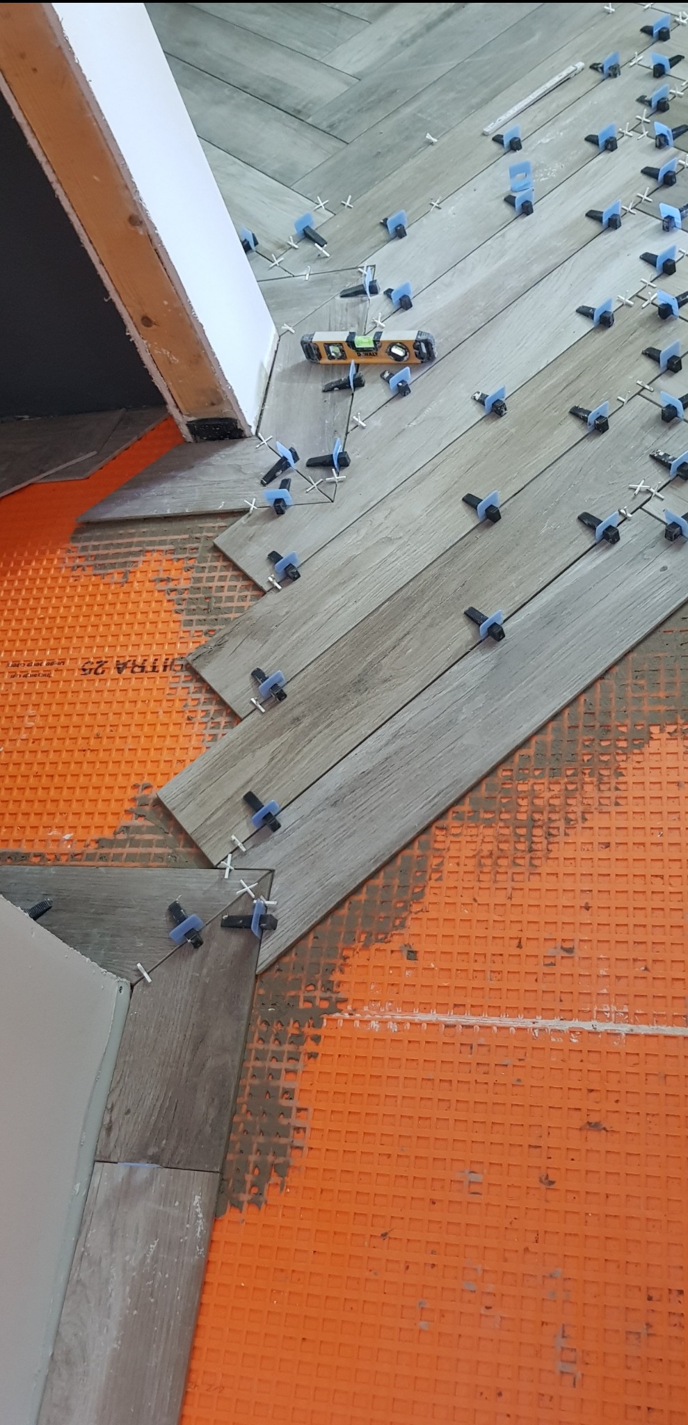 Układanie podłogi z płytek imitujących drewno w jodełkę, z użyciem poziomicy i systemu poziomowania płytek na pomarańczowej macie izolacyjnej.