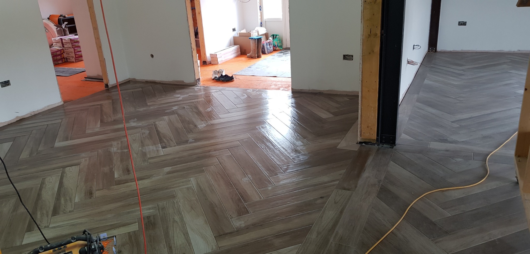 Ułożona podłoga z płytek imitujących drewno w jodełkę w trakcie prac wykończeniowych, widoczne narzędzia i kable, w tle materiały budowlane.