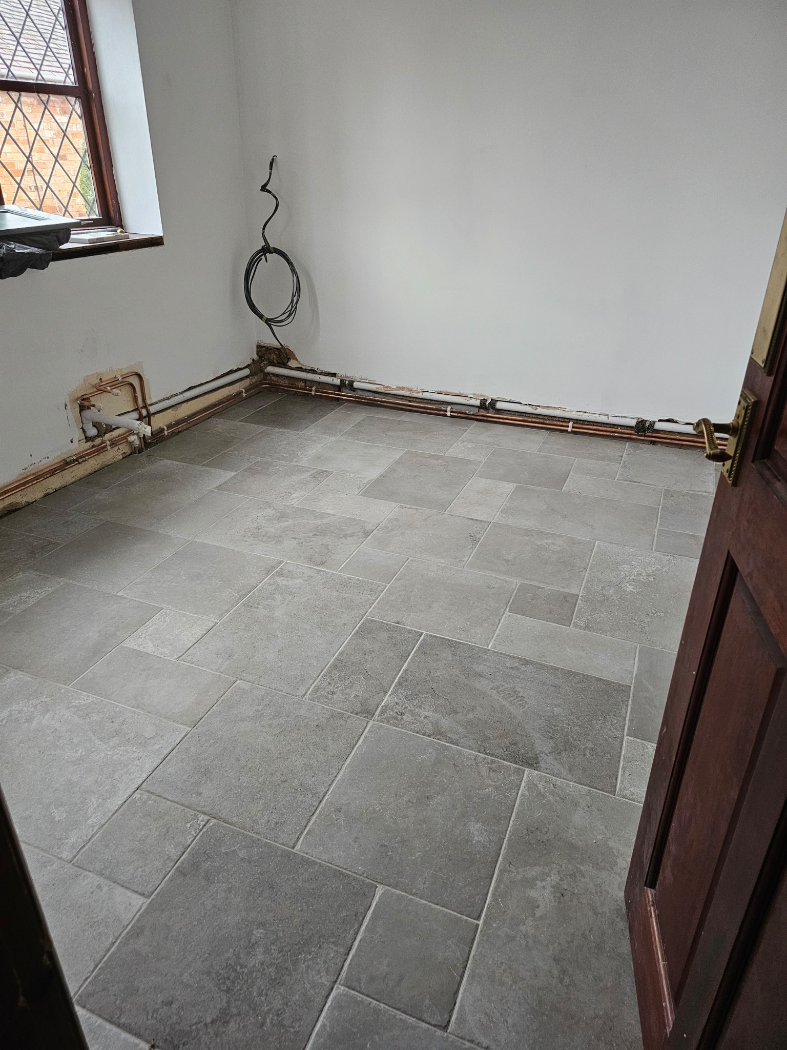 Wykończona podłoga z szarych płytek w pomieszczeniu w trakcie prac instalacyjnych; widoczne rury miedziane przy ścianie i przewód elektryczny zwisający ze ściany, okno z szybami w kratkę.