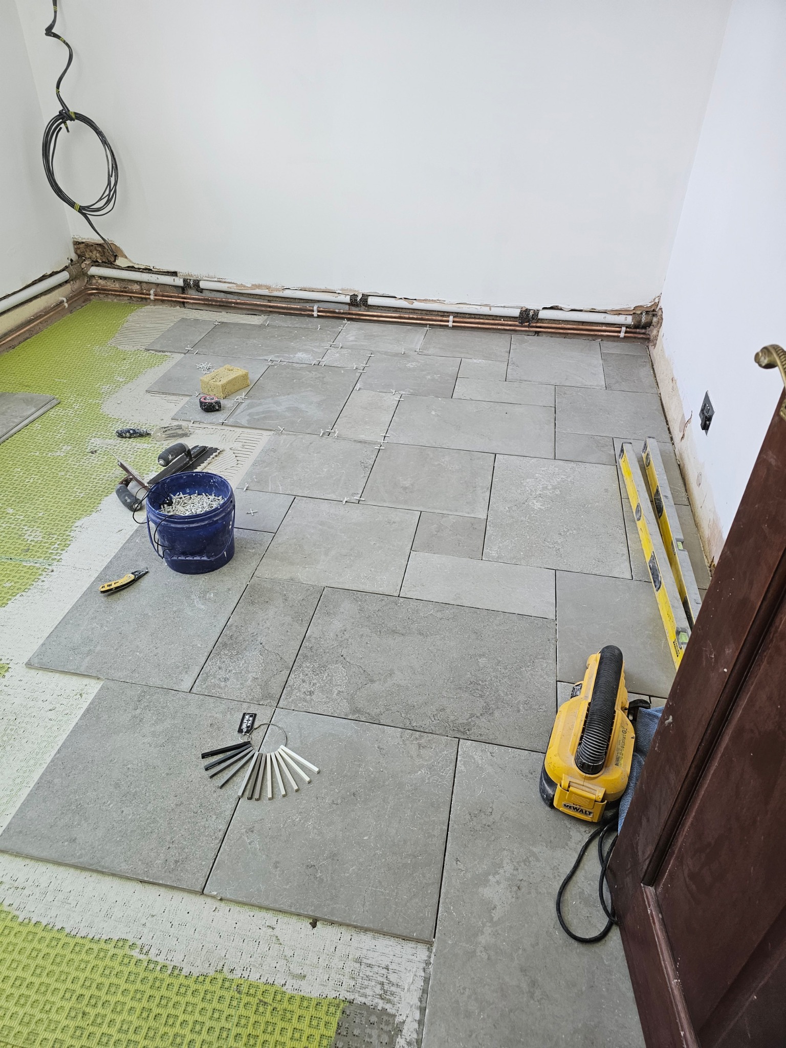 Układanie szarych płytek podłogowych w trakcie prac wykończeniowych, widoczny poziomica, wiadro z krzyżykami dystansowymi, próbnik kolorów fug i szlifierka DeWalt przy futrynie.