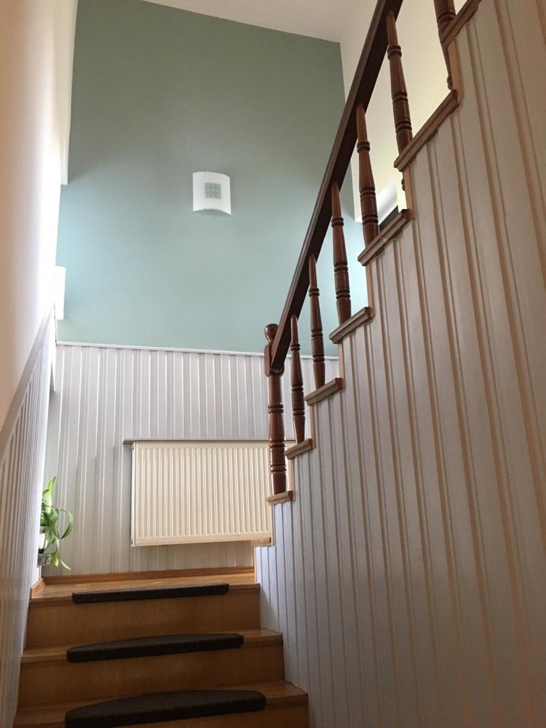 Klatka schodowa z drewnianymi schodami z czarnymi nakładkami, białą boazerią ścienną, pomalowaną na pastelowy zielony kolor górną częścią ściany i drewnianą balustradą z ciemnego drewna.