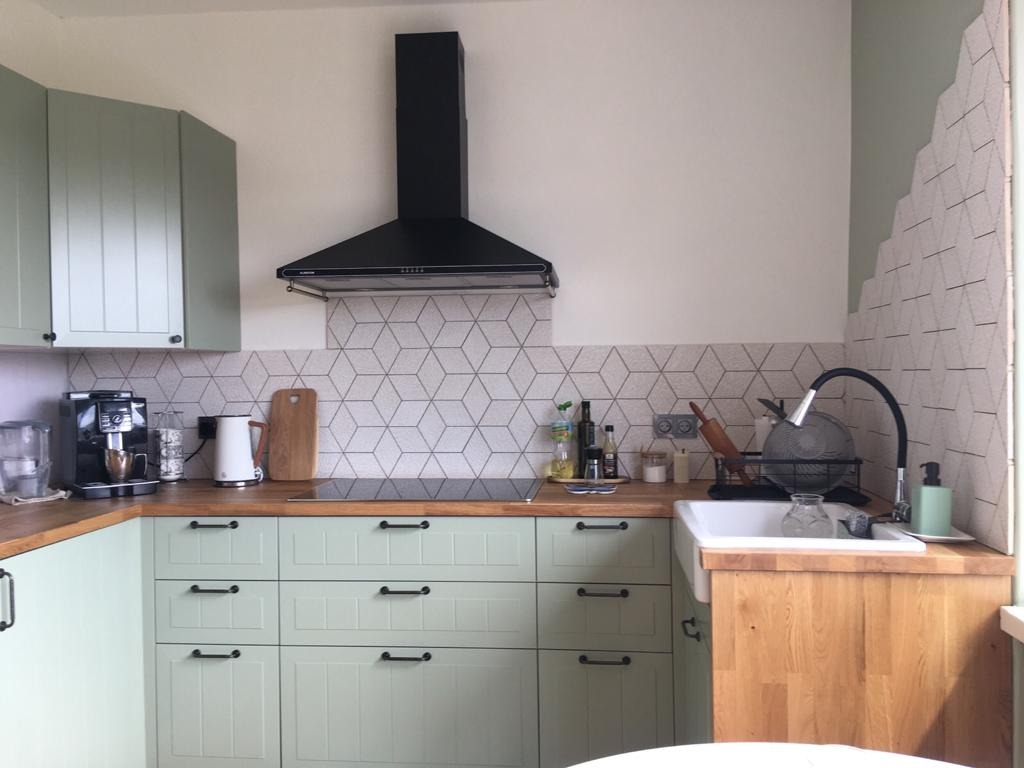 Jasnozielona kuchnia z drewnianym blatem, czarnym okapem i geometrycznymi płytkami na ścianie, widok na zlew z ociekaczem i akcesoriami kuchennymi.