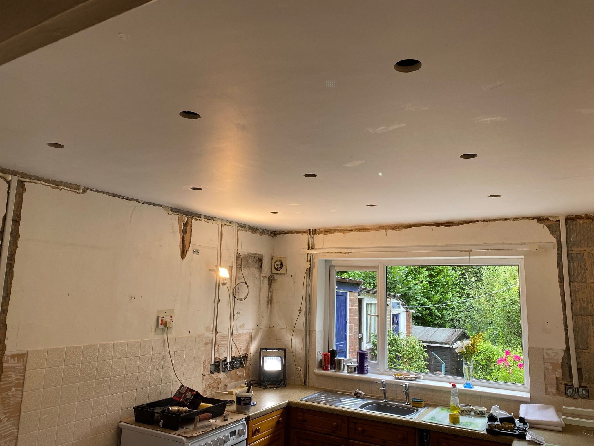 Kuchnia w trakcie demontażu: usunięte płytki, odkryte kable, sufit z wywierconymi otworami na oświetlenie, widok na zielony ogród przez okno.