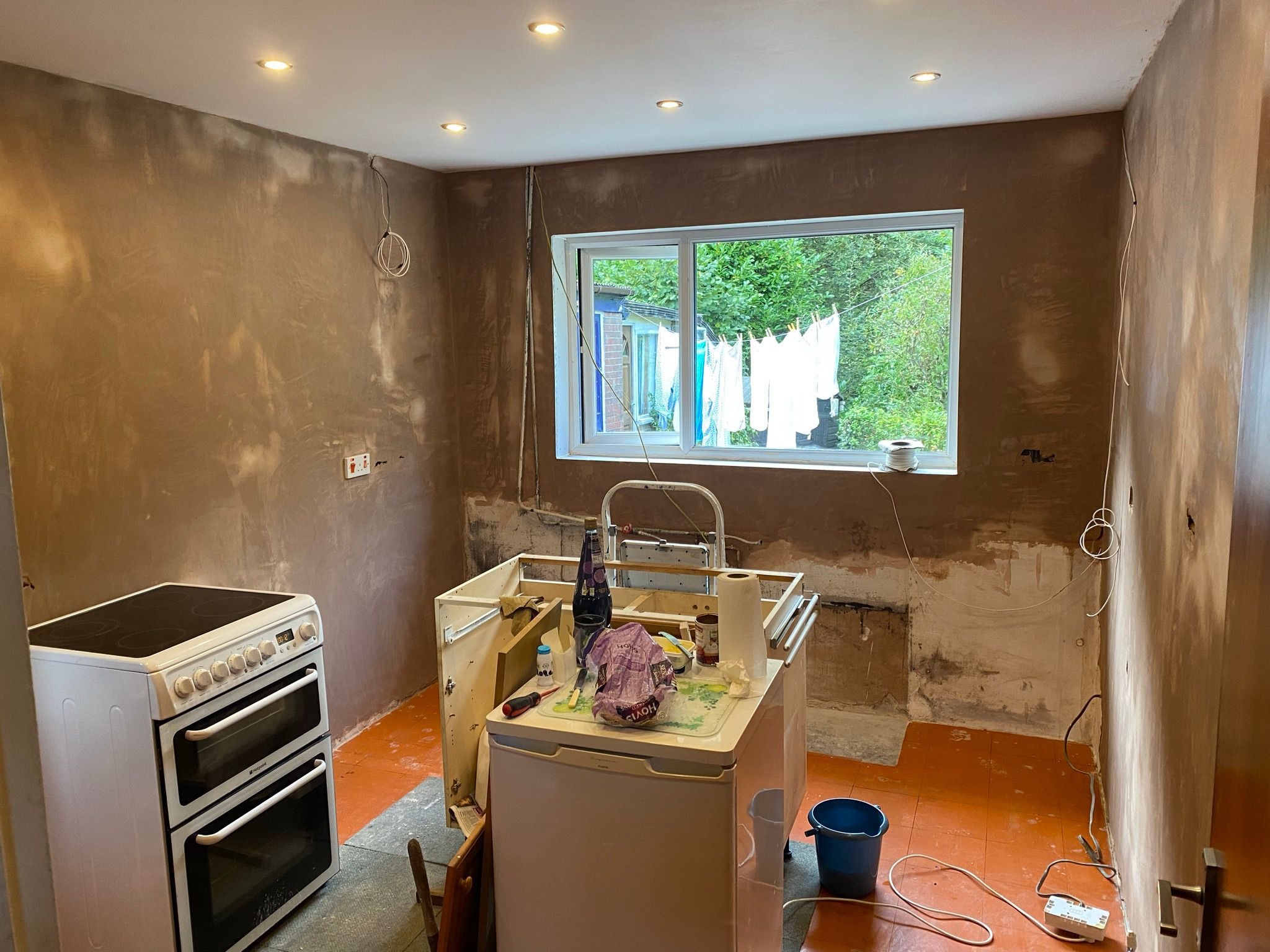 Kuchnia w trakcie szpachlowania ścian, widoczny stary piec, fragment szafki kuchennej, okno z suszącym się praniem na zewnątrz