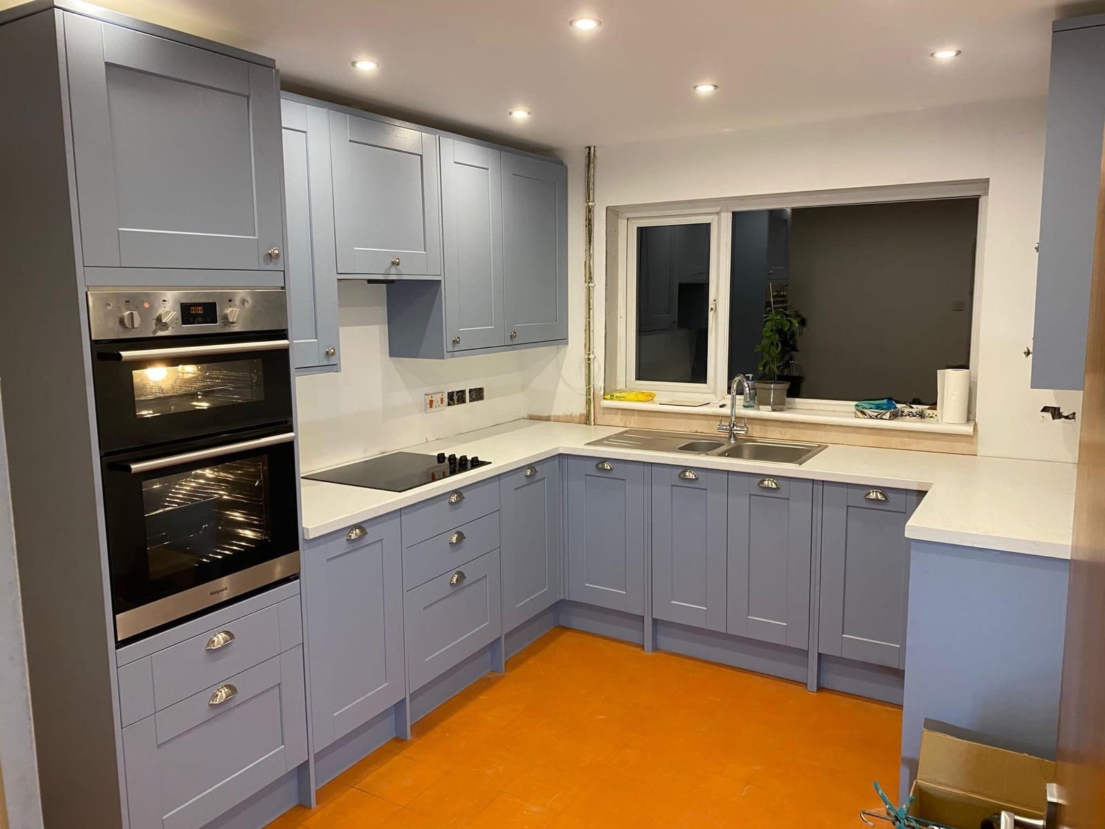 Kuchenna zabudowa w odcieniach szarości z białym blatem, dwukomorowy zlew pod oknem, płyta indukcyjna i piekarnik w zabudowie, pomarańczowa podłoga, widoczne gniazdka elektryczne.