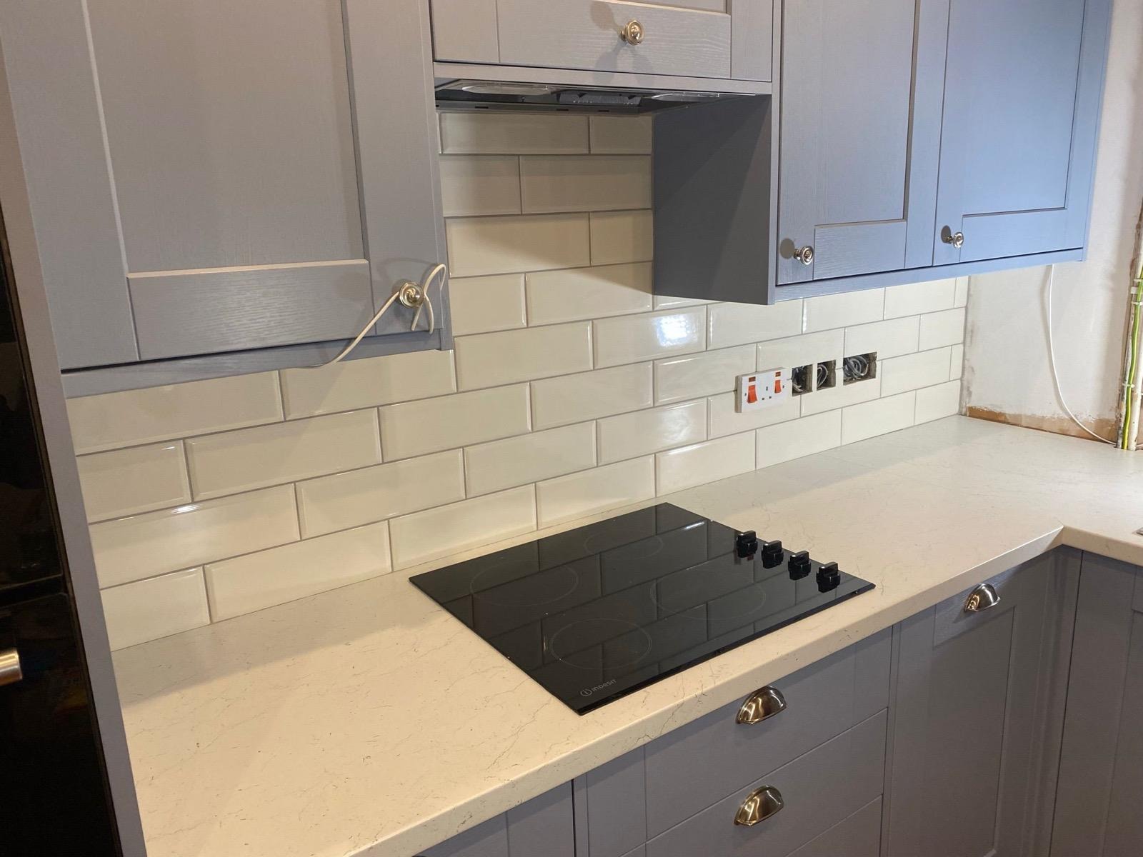 Kuchnia w trakcie wykończenia: szare szafki z frezowanymi frontami, białe płytki ułożone horyzontalnie, czarna płyta indukcyjna, widoczne gniazdka elektryczne bez osprzętu i wystające przewody