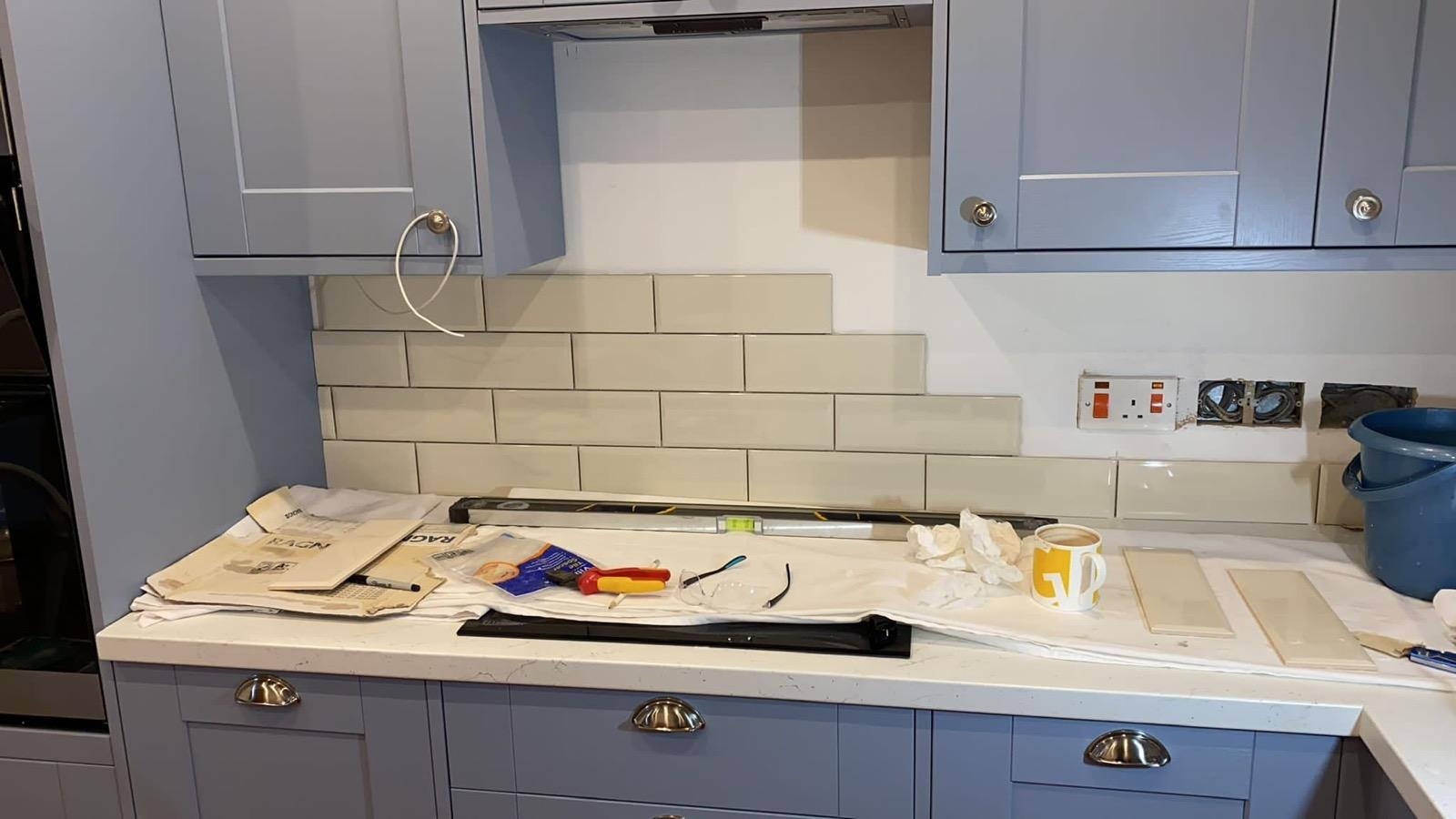 Układanie beżowych, prostokątnych płytek na ścianie w kuchni, widoczne puszki elektryczne, poziomica, narzędzia glazurnicze i kubek z kawą na blacie.