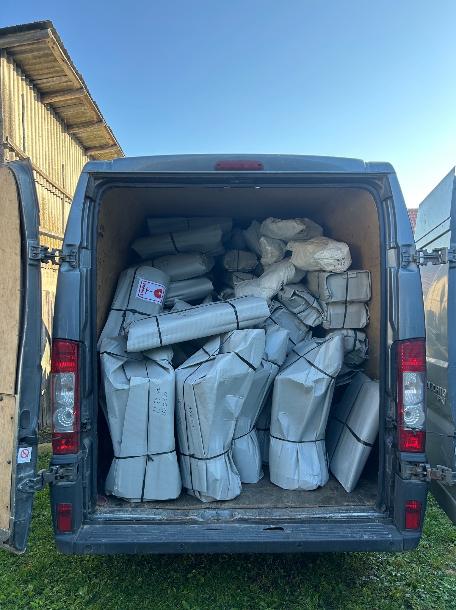 Przestrzeń ładunkowa busa wypełniona po brzegi paczkami owiniętymi szarym papierem i związanymi czarnym sznurkiem. Na niektórych paczkach widoczne napisy i naklejki 'Fragile'.