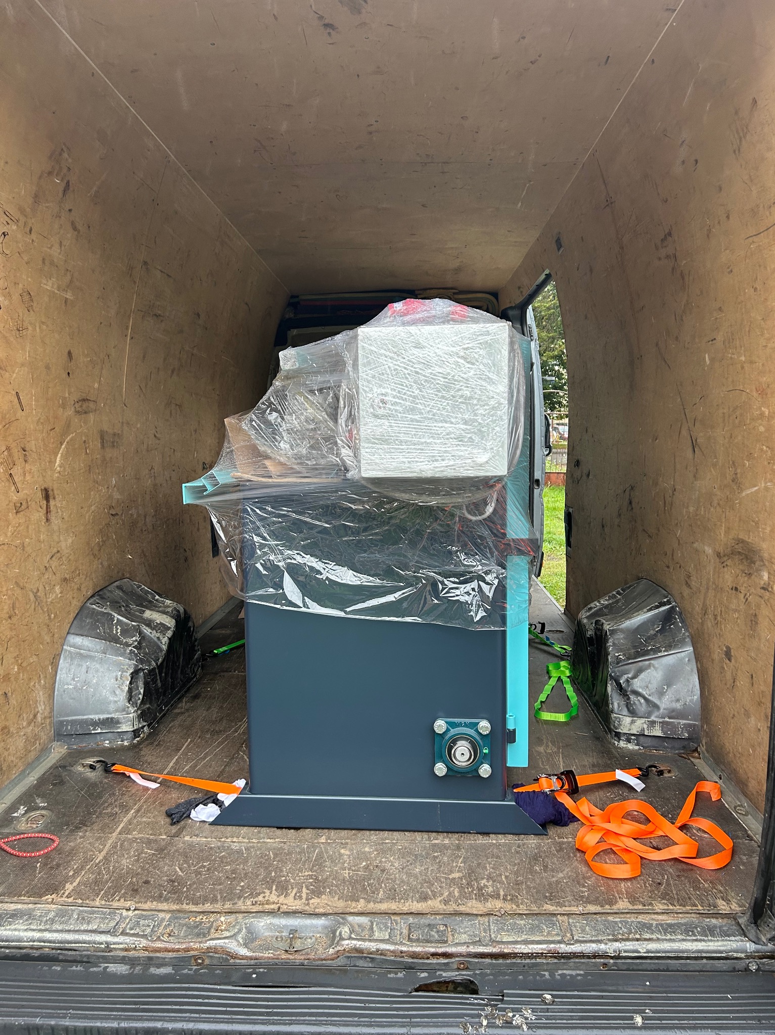 Maszyna owinięta folią stretch, zabezpieczona pasami transportowymi w przestrzeni ładunkowej busa. Widoczne elementy mocujące i ślady użytkowania na podłodze.