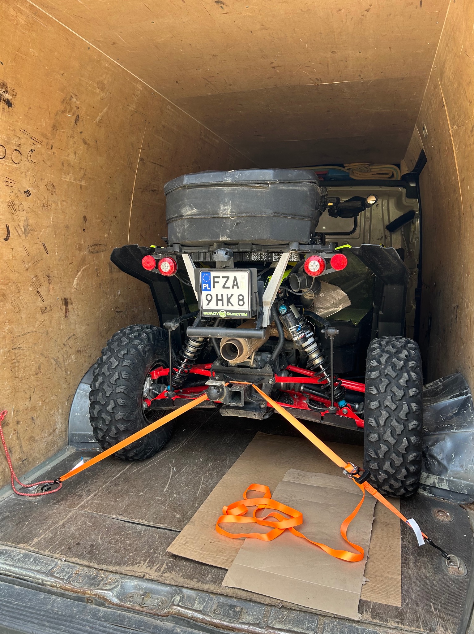 ATV (quad) zabezpieczony pomarańczowymi pasami wewnątrz skrzyni ładunkowej busa. Widoczna tylna część pojazdu i tablica rejestracyjna.
