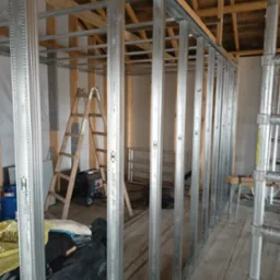 Konstrukcja ścian działowych z metalowych profili, widoczne drewniane elementy konstrukcyjne dachu, drabina i agregat prądotw&oacute;rczy w tle.