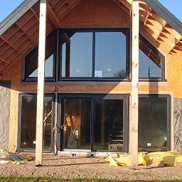 Predator Konstrukcje Tomasz Zieliński Usługi Ogólnobudowlane - Dom szkieletowy w trakcie budowy z widoczną konstrukcją drewnianą, czarnymi oknami i częściowo ocieploną elewacją, na pierwszym planie trawa i ziemia.