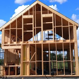 Predator Konstrukcje Tomasz Zieliński Usługi Ogólnobudowlane - Surowy szkielet dwukondygnacyjnego domu z drewna na fundamencie, z widocznymi elementami konstrukcyjnymi i fragmentem niebieskiego nieba w tle.