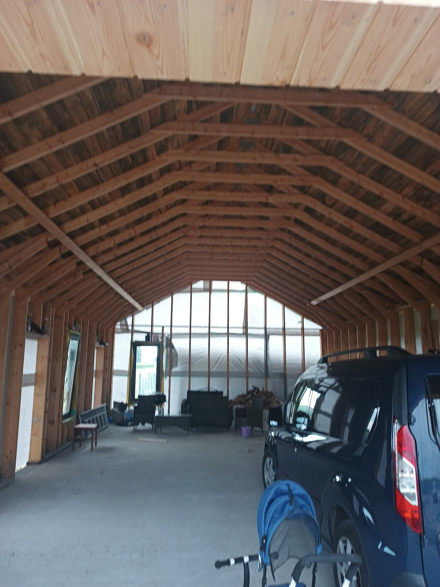 Wnętrze surowego garażu z drewnianą konstrukcją dachu, widoczne elementy domu szkieletowego i samochód oraz wózek dziecięcy w tle.