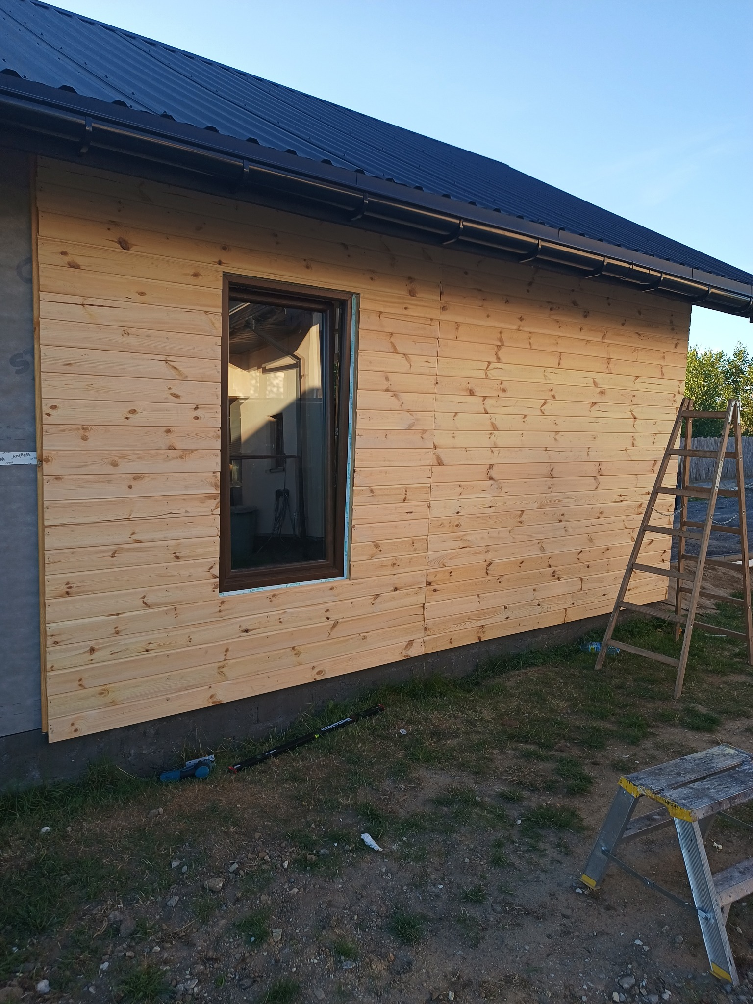 Fragment ściany zewnętrznej domu w trakcie budowy, pokrytej drewnianą boazerią, z widocznym oknem, czarnym dachem i drabiną opartą o ścianę.