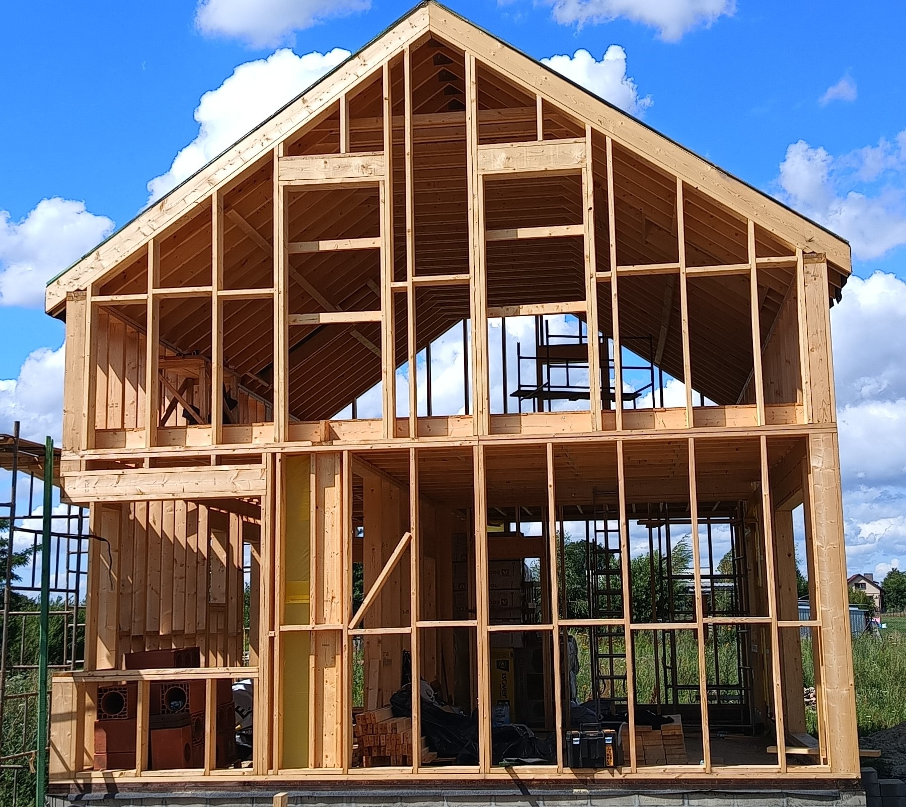Surowy szkielet dwukondygnacyjnego domu z drewna na fundamencie, z widocznymi elementami konstrukcyjnymi i fragmentem niebieskiego nieba w tle.
