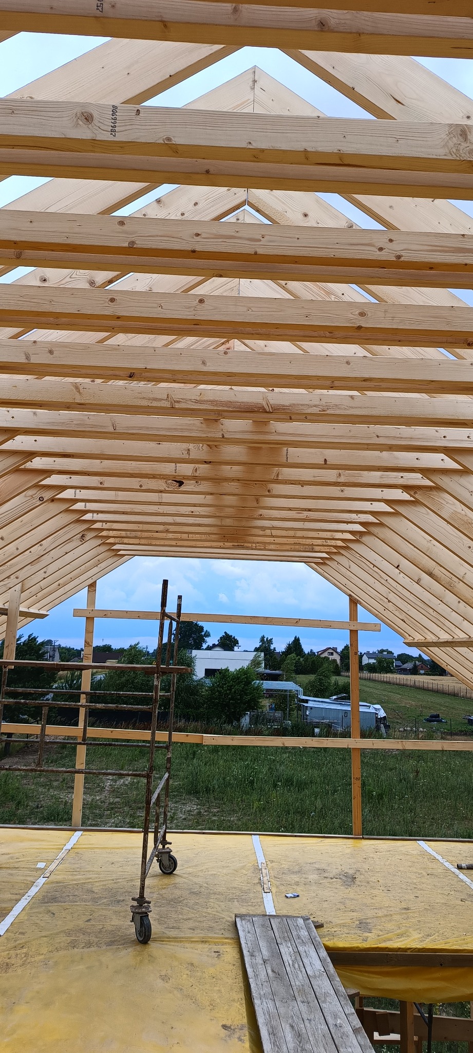 Konstrukcja drewniana dachu domu szkieletowego w trakcie budowy, widok z wnętrza, z żółtą folią izolacyjną na podłodze i krajobrazem w tle.