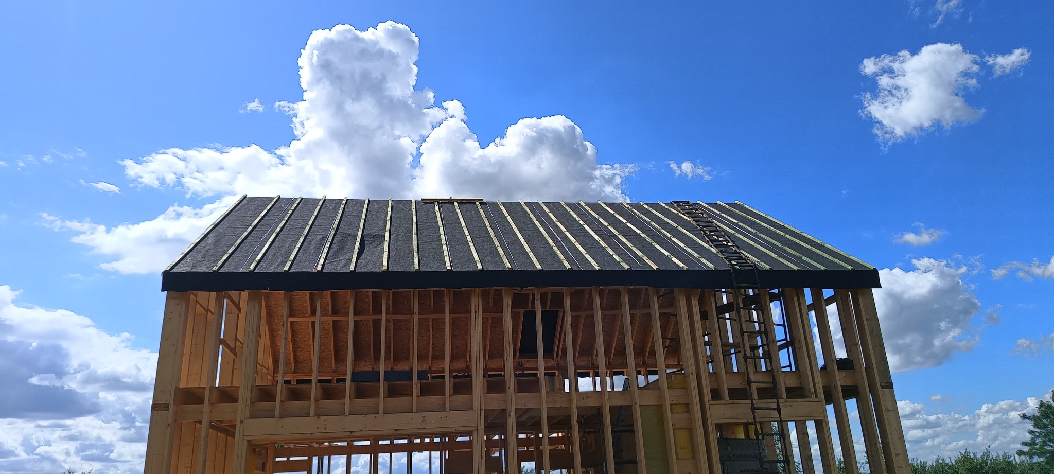 Konstrukcja domu szkieletowego z widocznym dachem w trakcie budowy, na tle błękitnego nieba z chmurami. Prace budowlane w Wierzbicy.