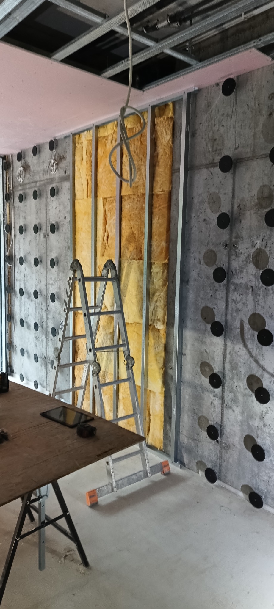 Ocieplanie ściany wełną mineralną w konstrukcji szkieletowej. Widoczna drabina, płyta karton-gips na suficie i betonowa ściana z czarnymi mocowaniami.