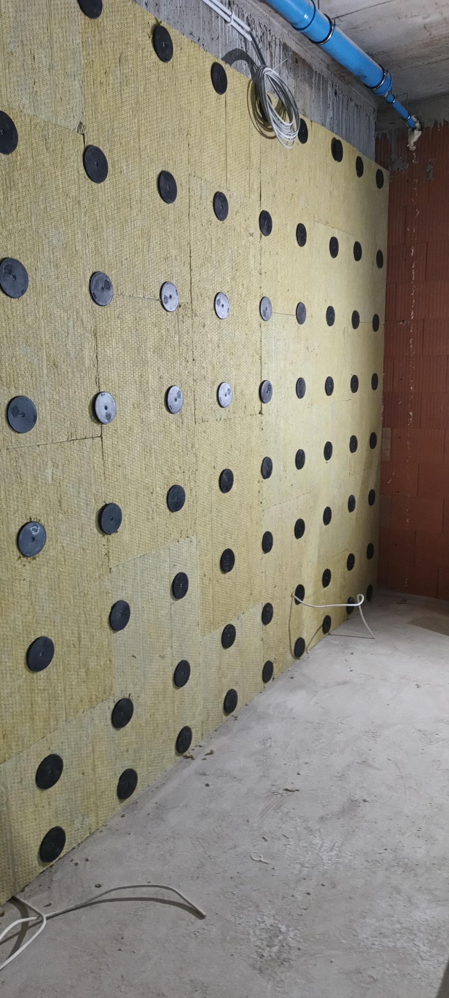 Żółta wełna mineralna przymocowana do ściany za pomocą czarnych uchwytów. Widoczne przewody i fragment ściany z cegły. Ocieplenie wewnętrzne w trakcie realizacji.