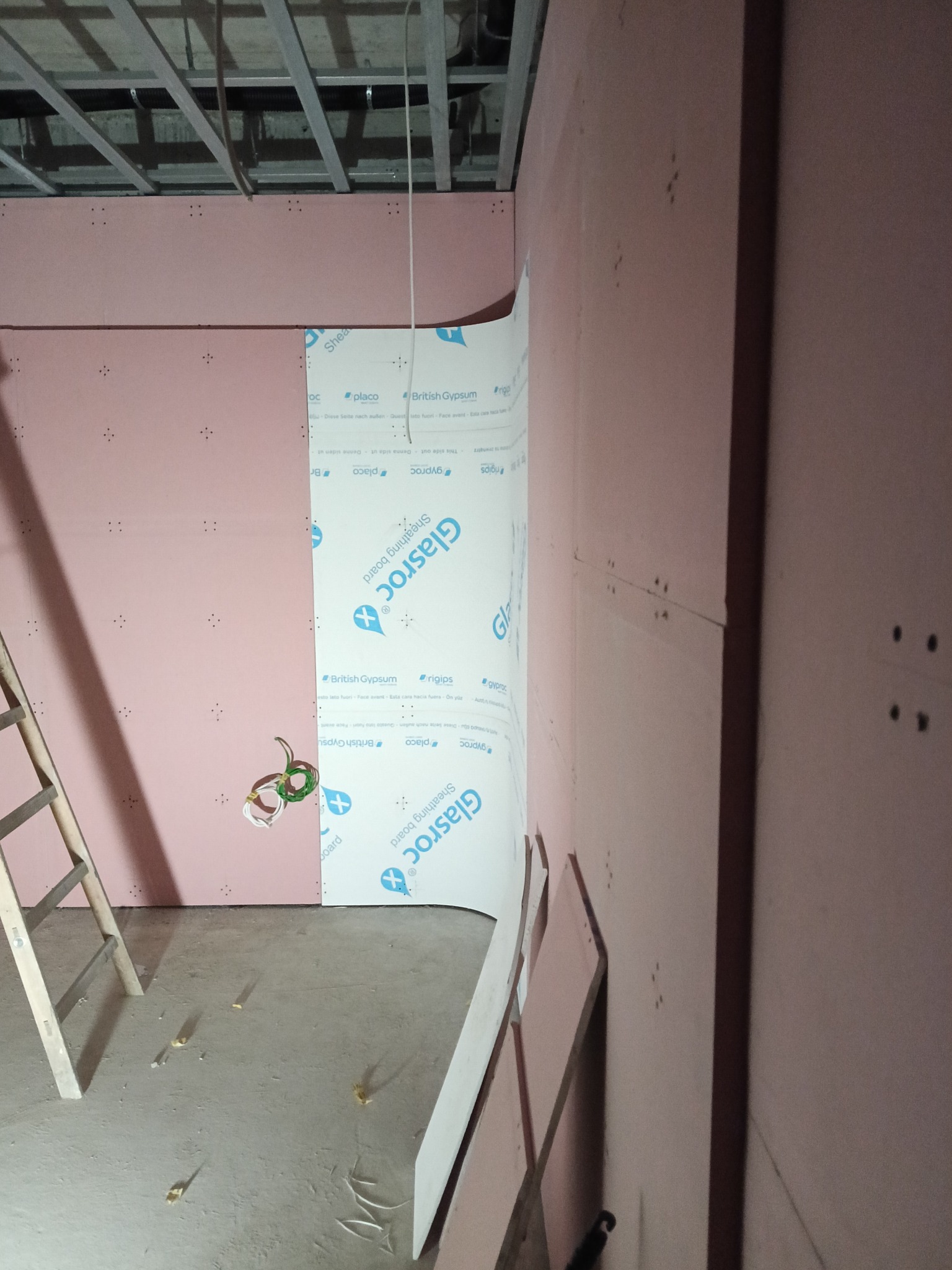 Montaż różowej płyty kartonowo-gipsowej Glasroc na ścianie z widoczną konstrukcją sufitu i fragmentem drabiny. Widoczne przewody elektryczne.