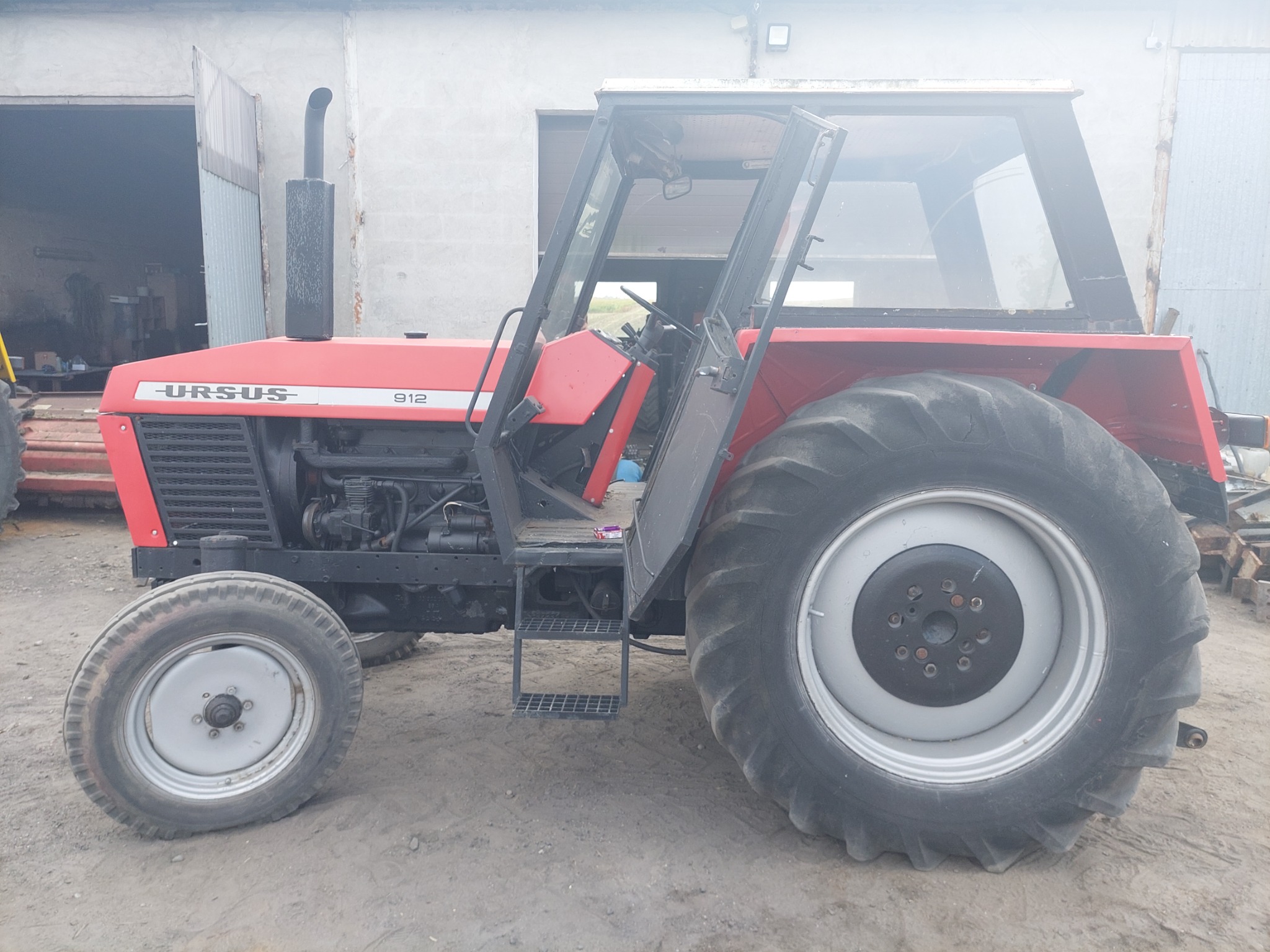 Czerwony ciągnik rolniczy Ursus 912 z otwartymi drzwiami kabiny, zaparkowany na żwirowym podłożu przed otwartą, szarą halą.