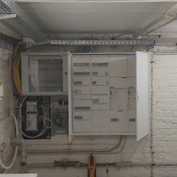 El-Smart Instalacje elektryczne GRZEGORZ GRALAK - Otwarta skrzynka elektryczna z widocznymi bezpiecznikami i przewodami w pomieszczeniu z pomalowanymi na biało ceglanymi ścianami i instalacją pod sufitem.