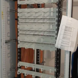 Rozdzielnica teleinformatyczna z pomarańczowymi kablami, listwami krosowniczymi i dokumentacją techniczną po prawej stronie.