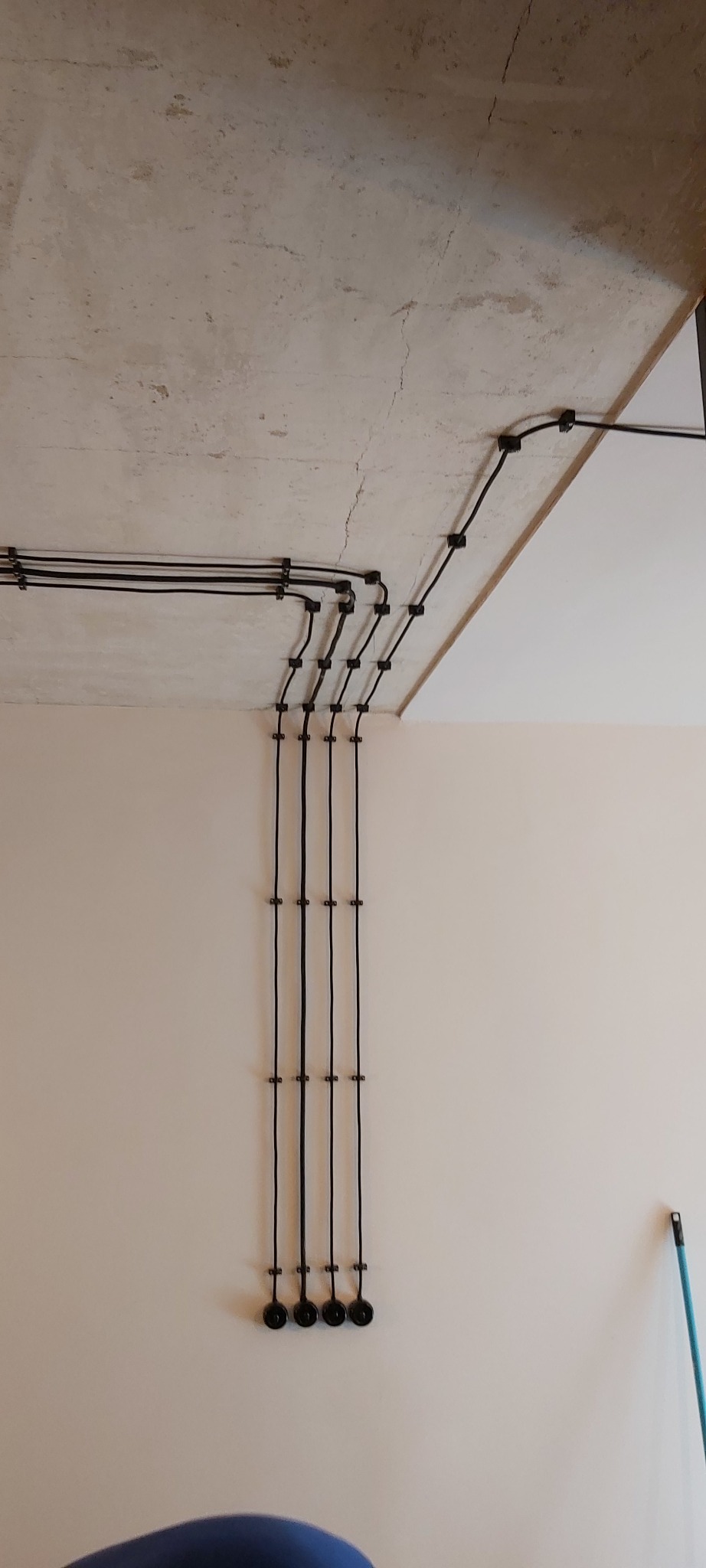 Cztery czarne przewody elektryczne prowadzone po powierzchni ściany i sufitu, przymocowane za pomocą uchwytów, widoczne puszki instalacyjne na dole ściany.