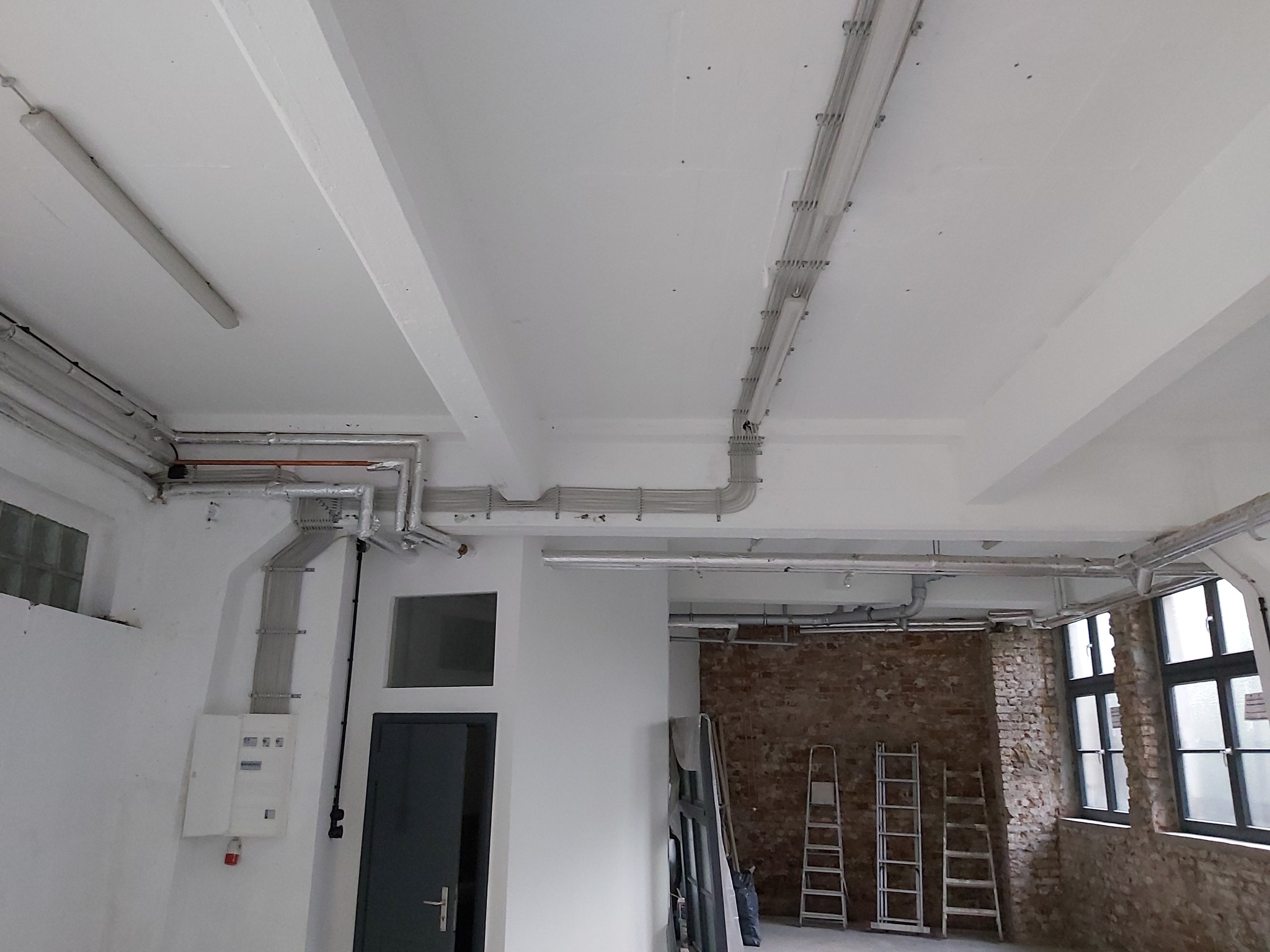 Białe wnętrze z widocznymi instalacjami elektrycznymi pod sufitem, koryta kablowe prowadzone wzdłuż sufitu i ścian, rozdzielnica na ścianie, surowe wykończenie z fragmentami odsłoniętej cegły w tle.