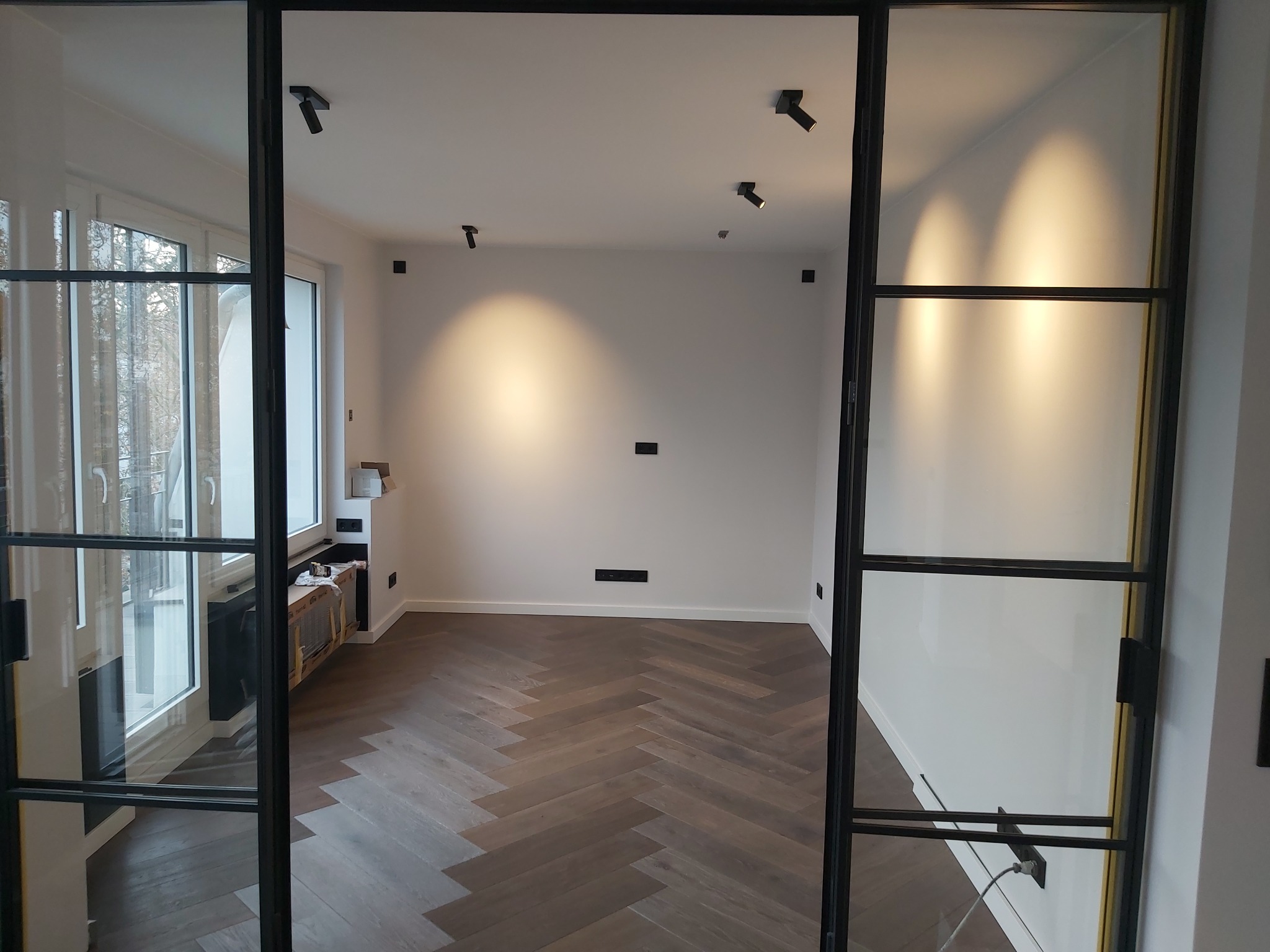 Nowoczesne wnętrze z parkietem w jodełkę, oświetleniem punktowym na suficie i przeszklonymi drzwiami w czarnych ramach.