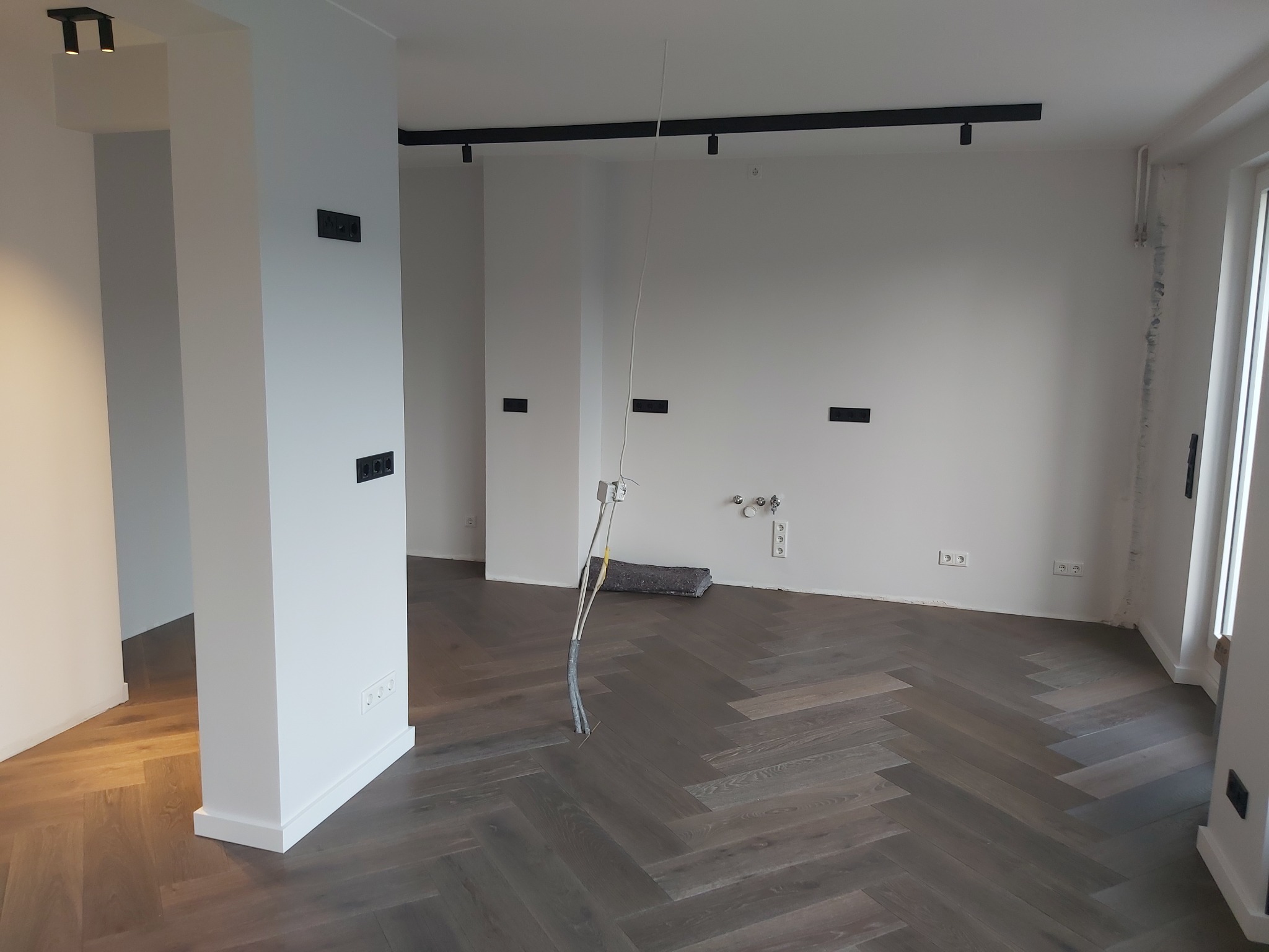 Wnętrze w trakcie remontu z parkietem w jodełkę, widoczne przewody wystające z podłogi, czarne gniazdka elektryczne na białych ścianach i szynoprzewody z reflektorami na suficie.